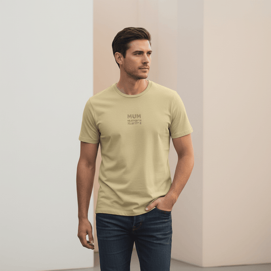 Male model wearing a beige Supima cotton T-shirt by Lilak with ‘MUM’ and Mumbai coordinates printed on the center chest, standing in a modern softly lit studio environment