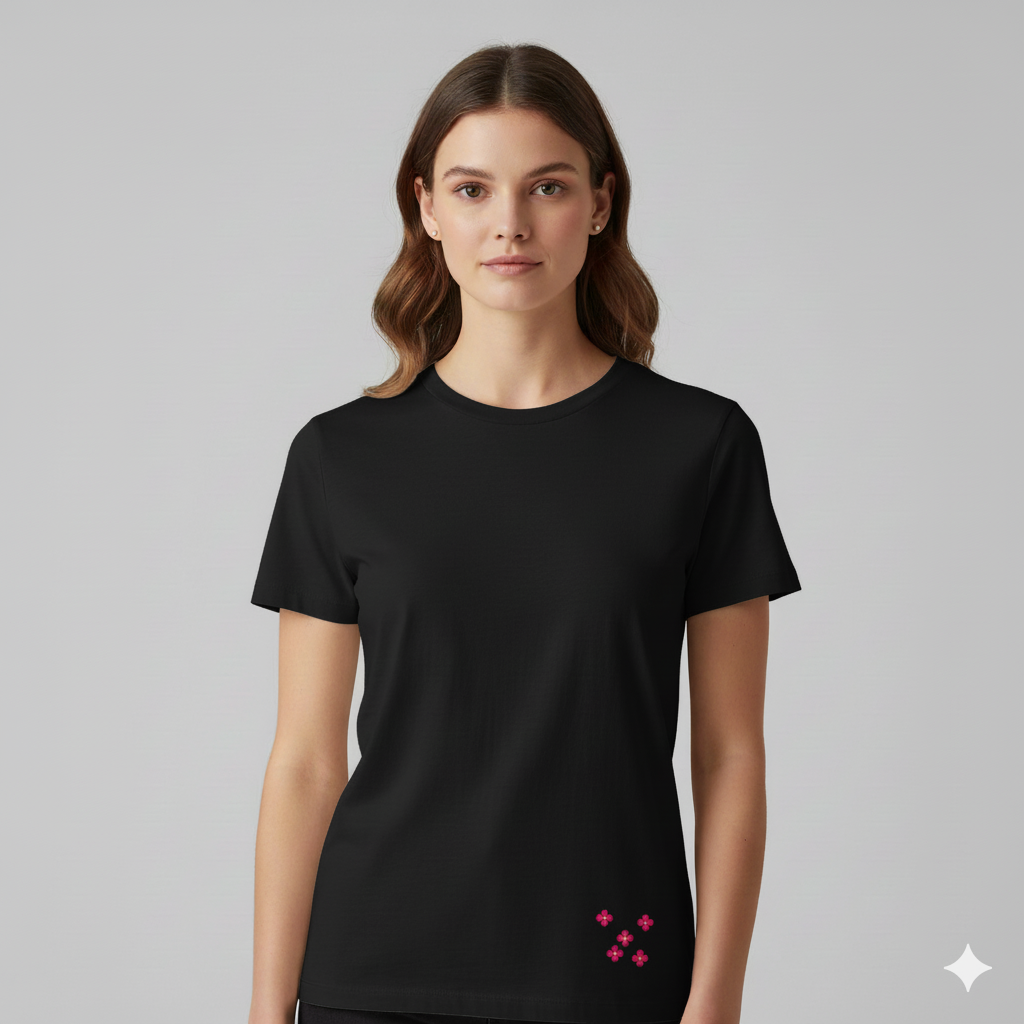 Female model wearing a Black Supima cotton T-shirt by Lilak, featuring a small pink floral cluster printed near the bottom left hem, photographed against a plain light grey studio background