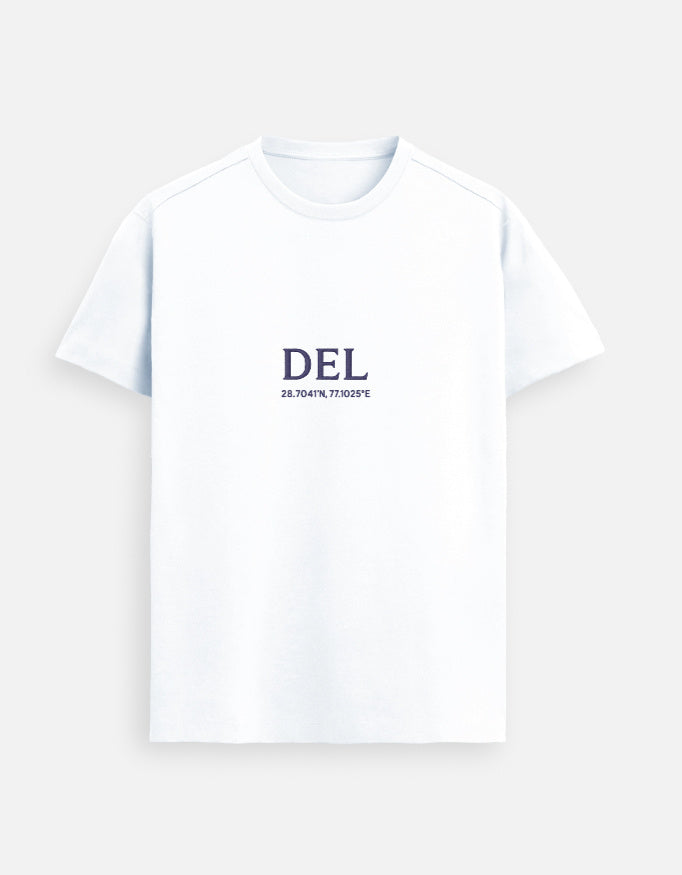 Male model standing indoors wearing a White Lilak Supima cotton T-shirt with ‘DEL’ and geographic coordinates printed on the chest, photographed in soft natural light with a plant and wooden furniture in the background