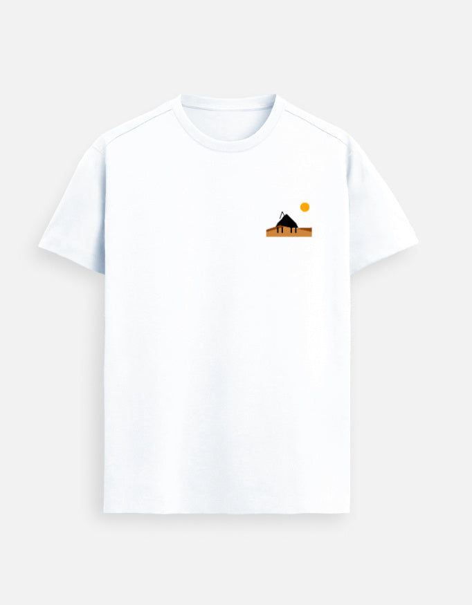 Male model standing in a softly lit studio wearing a White Supima cotton T-shirt by Lilak, featuring a minimal sunset hut graphic on the left chest