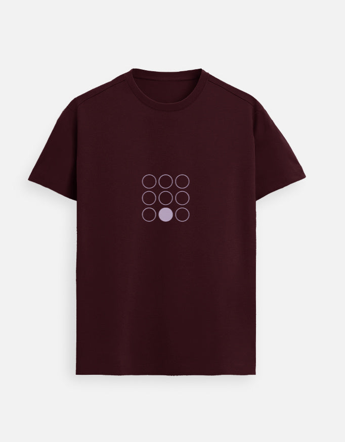 Indian female model wearing a Maroon Supima cotton T-shirt by Lilak, featuring a minimal lavender geometric circle design on the center chest. She stands in a softly lit studio with large window shadows in the background