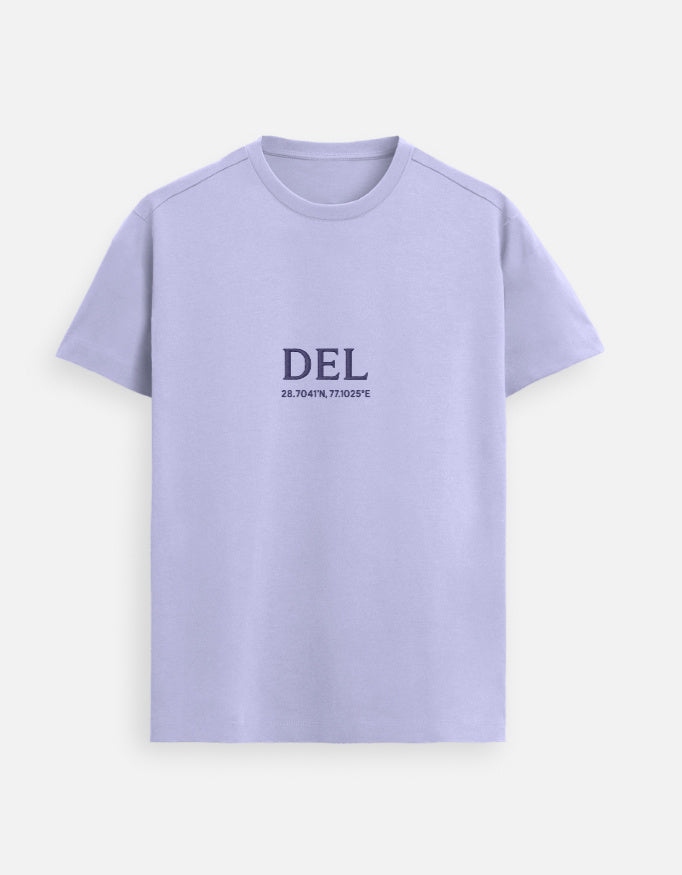 Male model standing indoors wearing a Lavender Lilak Supima cotton T-shirt with ‘DEL’ and geographic coordinates printed on the chest, photographed in soft natural light with a plant and wooden furniture in the background