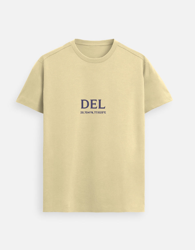 Male model standing indoors wearing a Beige Lilak Supima cotton T-shirt with ‘DEL’ and geographic coordinates printed on the chest, photographed in soft natural light with a plant and wooden furniture in the background