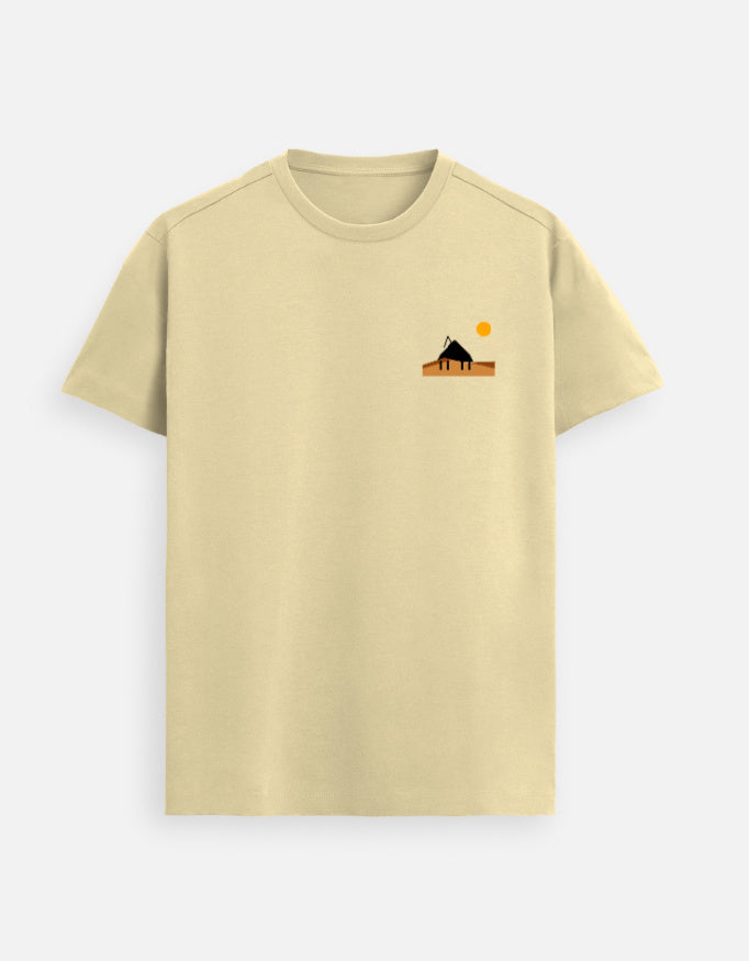 Male model standing in a softly lit studio wearing a Beige Supima cotton T-shirt by Lilak, featuring a minimal sunset hut graphic on the left chest