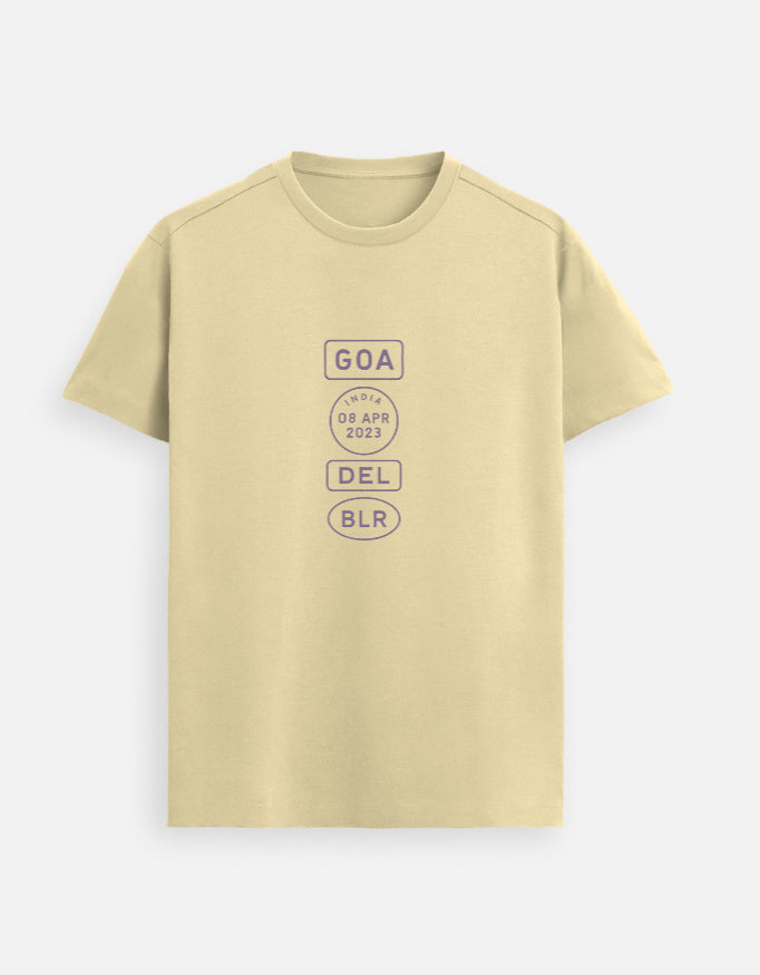 Indian male model wearing a Beige Supima cotton T-shirt by Lilak, featuring a minimalistic violet travel-stamp design reading GOA, INDIA 08 APR 2023, DEL, and BLR. Model stands against a textured grey studio background