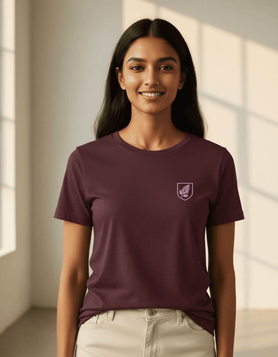 Female model wearing a Maroon Supima cotton T-shirt by Lilak featuring a small maroon botanical crest on the left chest, standing in a softly lit studio with large window shadows