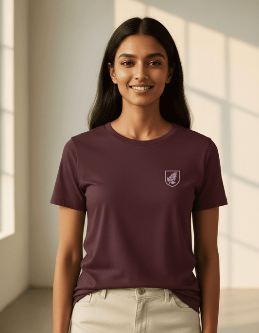 Female model wearing a Maroon Supima cotton T-shirt by Lilak featuring a small maroon botanical crest on the left chest, standing in a softly lit studio with large window shadows