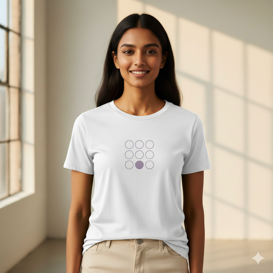 Indian female model wearing a White Supima cotton T-shirt by Lilak, featuring a minimal lavender geometric circle design on the center chest. She stands in a softly lit studio with large window shadows in the background
